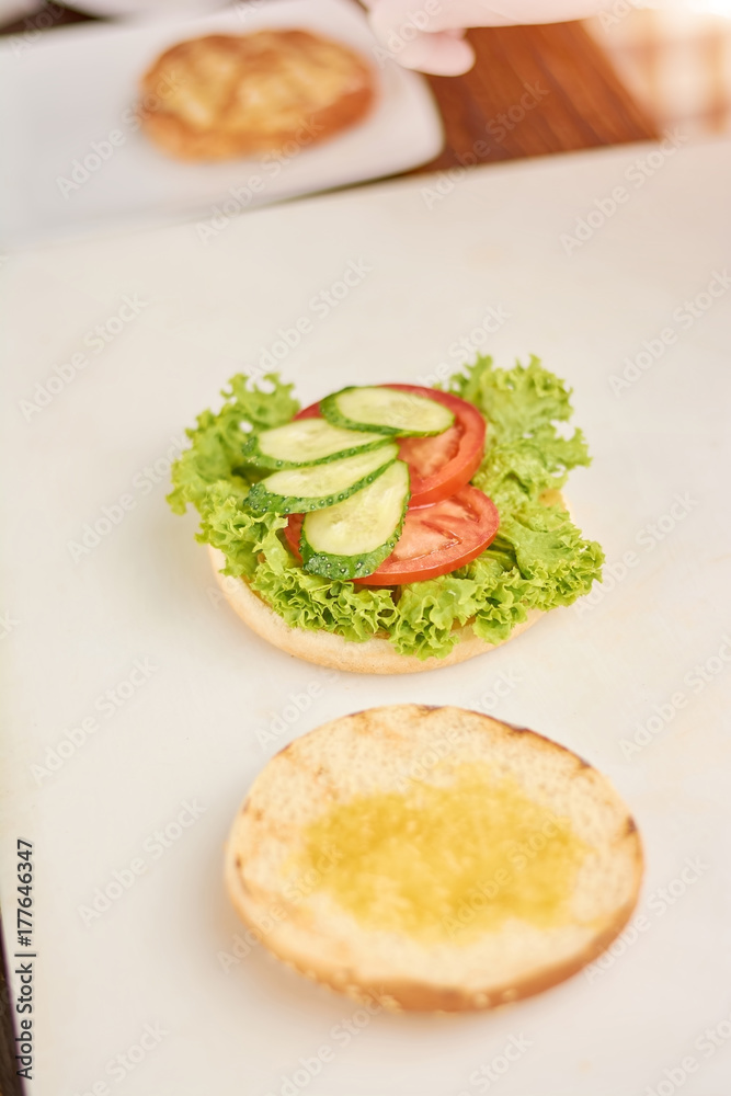 Cooking of hamburger at kitchen. Burger with lettuce, tomatoes and fresh cucumbers on table. Burger preparing at cafe. Cooking of hamburger at kitchen. Burger with lettuce, tomatoes and fresh cucumbers on table. Burger preparing at cafe.