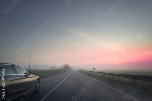 summer rural landscape with blue and red sky, fog and the road. sunrise. Dark car is moving to the sun