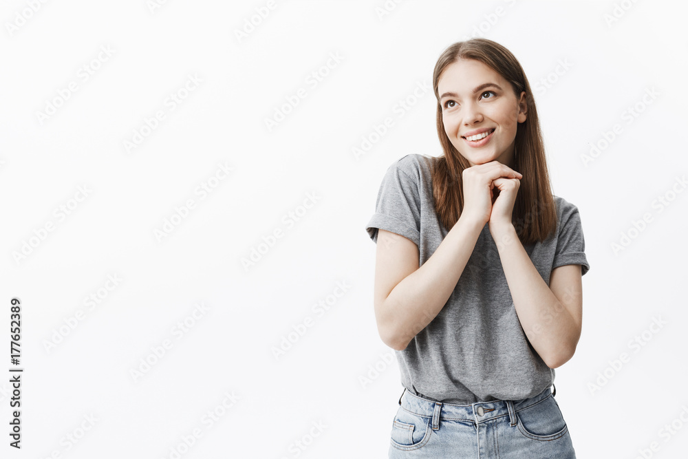 Close up of charming good-looking young student girl with dark hair and eyes in casual outfit smiling, looking aside with dreamy and happy face expression, holding hands together under face. Close up of charming good-looking young student girl with dark hair and eyes in casual outfit smiling, looking aside with dreamy and happy face expression, holding hands together under face.