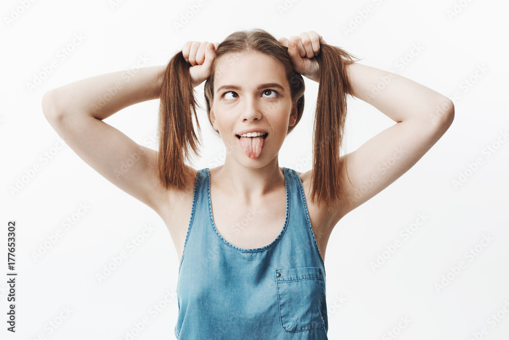 Positive emotions and expressions. Young good-looking skinny caucasian student girl holding hair in hands, showing tongue, mowing eyes, making funny expressions, having fun posing for university Positive emotions and expressions. Young good-looking skinny caucasian student girl holding hair in hands, showing tongue, mowing eyes, making funny expressions, having fun posing for university