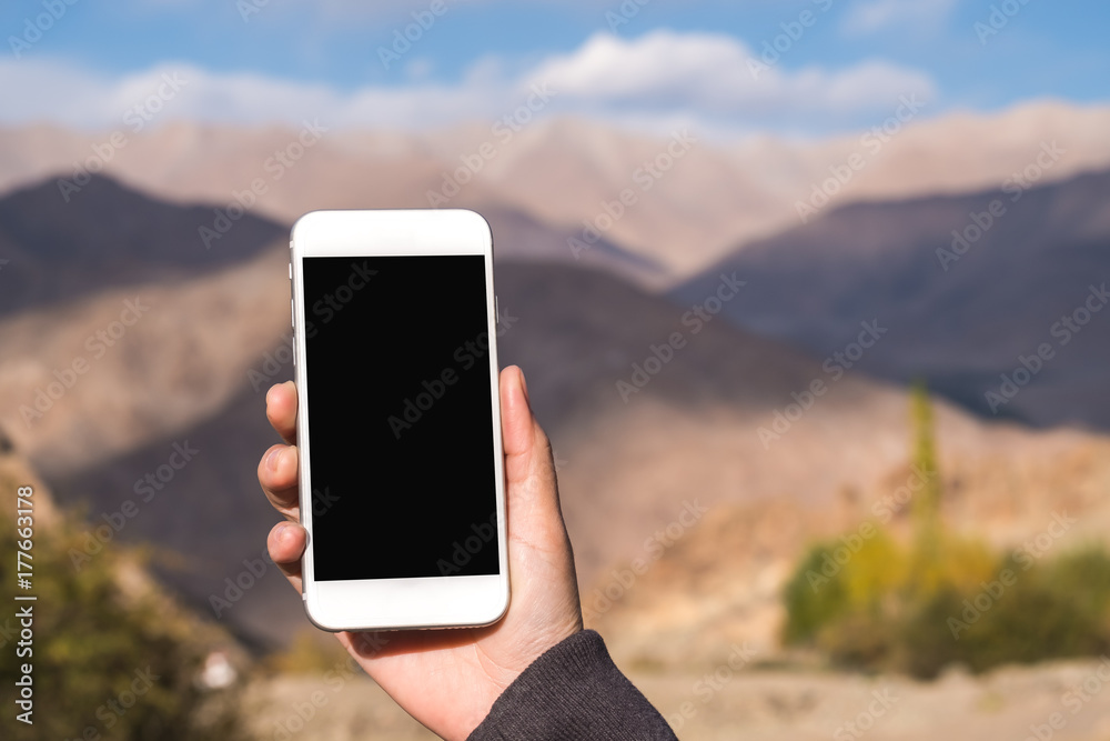 Mockup image of a hand holding and using white mobile phone with blank black screen while standing in front of mountains and blue sky Mockup image of a hand holding and using white mobile phone with blank black screen while standing in front of mountains and blue sky
