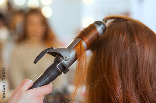 Hairdresser makes hairstyle girl with long red hair in a beauty salon. Create curls with curling irons. Professional hair care.