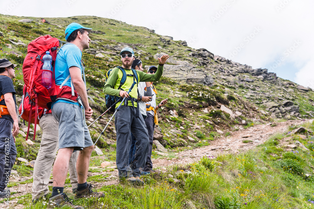 A group of hikers with backpacks and tracking sticks rest and stands in the mountains listening to their guide