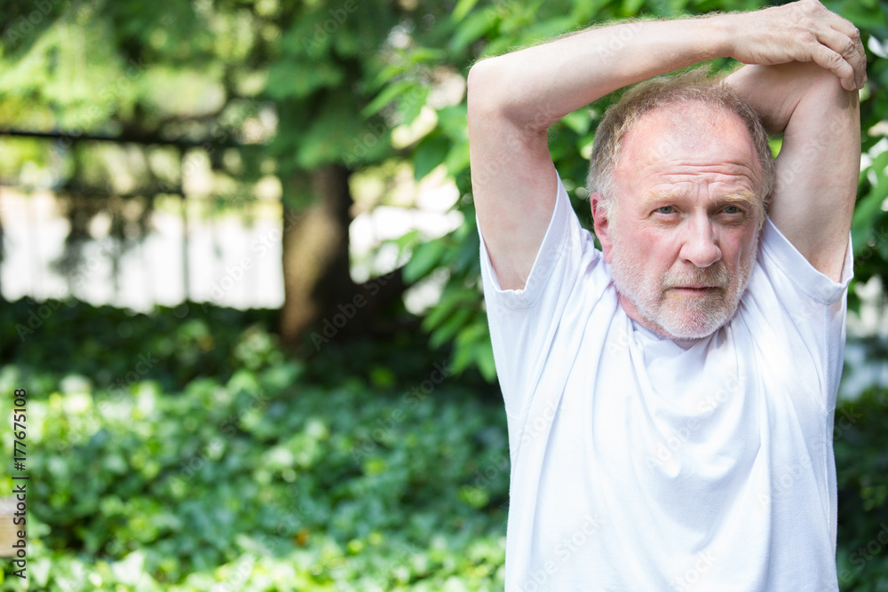 Obraz premium Closeup portrait, senior mature man in white shirt, stretching arms, isolated green trees background. Warming up