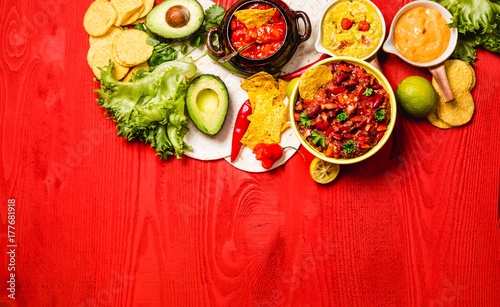 Vegetarian Mexican food concept: refried black and red beans. guacamole, salsa, chili, tortilla chips and fresh ingredients over vintage red rustic wooden background. Top view