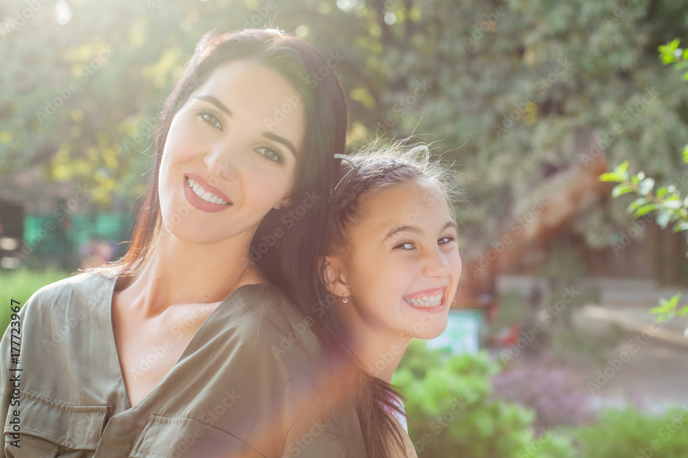 Young mom and daughter sitting back to back in park on sunny day and laughting Young mom and daughter sitting back to back in park on sunny day and laughting