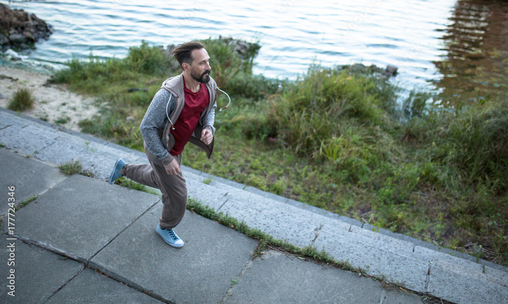 Man doing cardio workout outdoors. Beardy male jogging on river bank.