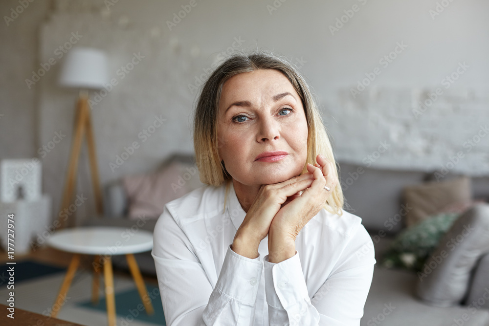 Head and shoulders of beautiful graceful senior woman with healthy wrinkled skin and loose thick hair having rest indoors, posing in modern interior, looking at camera with thoughtful face expression Head and shoulders of beautiful graceful senior woman with healthy wrinkled skin and loose thick hair having rest indoors, posing in modern interior, looking at camera with thoughtful face expression