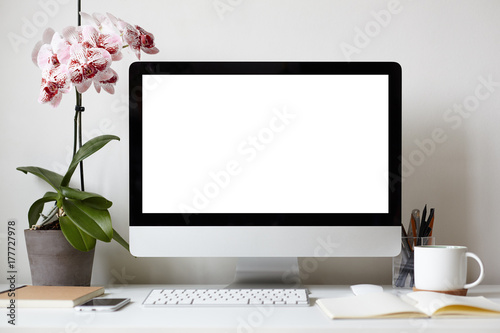 Picture of modern personal computer with blank white copy space screen resting on desk with keyboard, orchid in pot, mug, cellular phone, copybooks and stationery items. Mock up of workplace