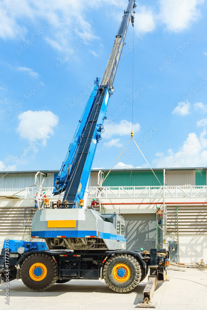 blue hydraulic truck crane standing on a construction site under construction. A worker is standing on a scissor lift at a suitable height to help positioning it.