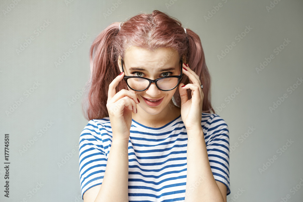 Picture of charming playful pink haired teenage girl with two ponytails and bright make up posing at grey wall peeking at camera over her stylish eyeglasses. People and optics advertisement