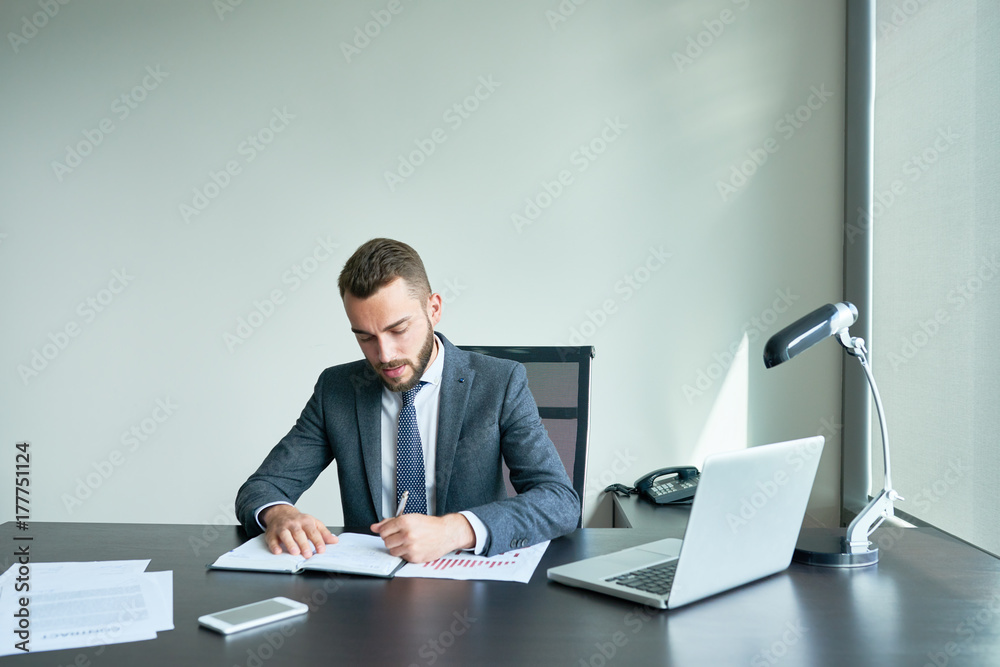 Waist-up portrait of confident young entrepreneur in classical suit taking necessary notes while preparing for important negotiations with business partners, interior of modern office on background Waist-up portrait of confident young entrepreneur in classical suit taking necessary notes while preparing for important negotiations with business partners, interior of modern office on background