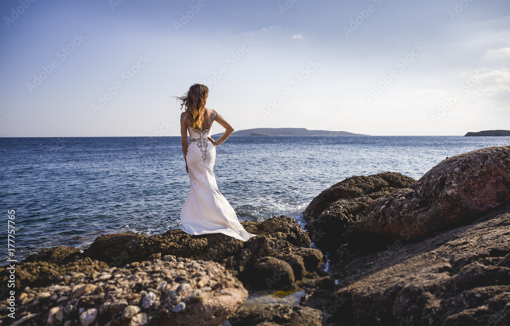 Bride standing on rocks on a sea shore and looking at water