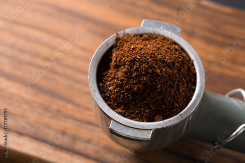 Pile of freshly roasted ground coffee in metal filter of the espresso machine on wooden surface ready to go to espresso machine with empty glass waiting