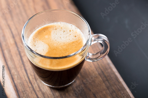 Glass of americano espresso fresh made dark black strong coffee on wooden board table, thick froth foam in cafe, morning refreshing hot drink caffeine, copy space for text