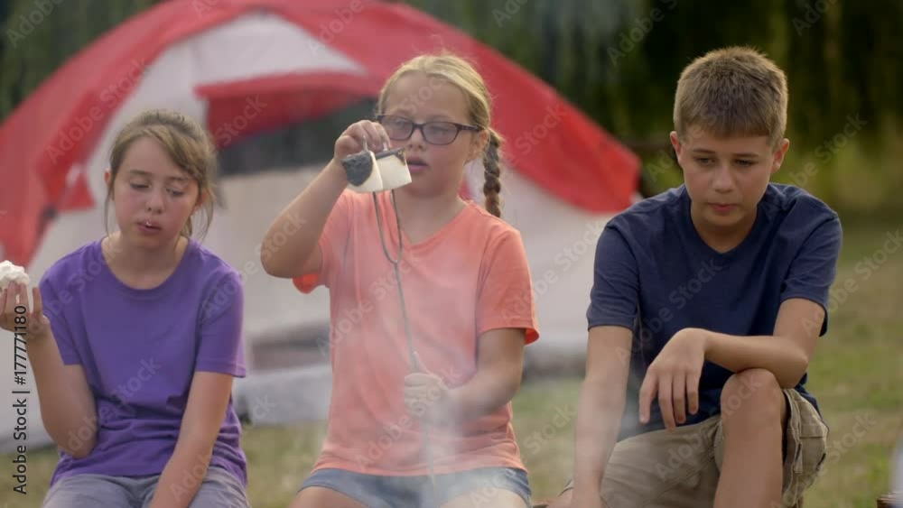 Girl pulls burned bits off toasted marshmallows on skewers beside an open fire at summer camp. Medium shot with two other campers, cooking and eating. Slow motion 4K recorded at 60fps.
