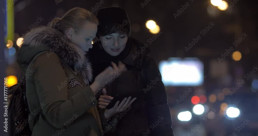Two young women discussing something using digital tablet while waiting for a bus at public transport stop in the evening Two young women discussing something using digital tablet while waiting for a bus at public transport stop in the evening