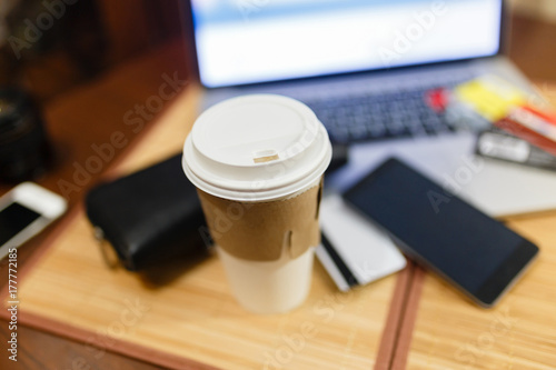 White paper Cup with burning coffee on the background of laptop and smartphone. Scattered Bank cards and a black purse. Work on the go.