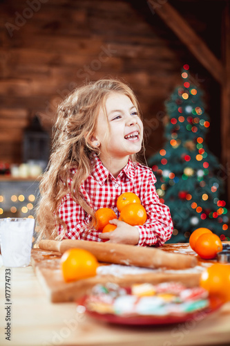 Happy little child, cute baby girl at table in home kitchen holds a lot of the orange and have fun in a Christmas interior with tree