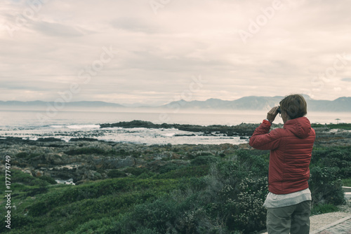 Tourist looking with binocular on the rocky coast line at De Kelders, South Africa, famous for whale watching. Winter season, cloudy and dramatic sky, toned image.