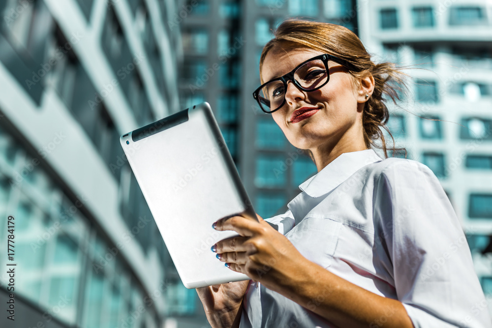 Young blonde caucasian businesswoman wears glasses and white blouse use tablet pc before the modern corporation building in the background Young blonde caucasian businesswoman wears glasses and white blouse use tablet pc before the modern corporation building in the background