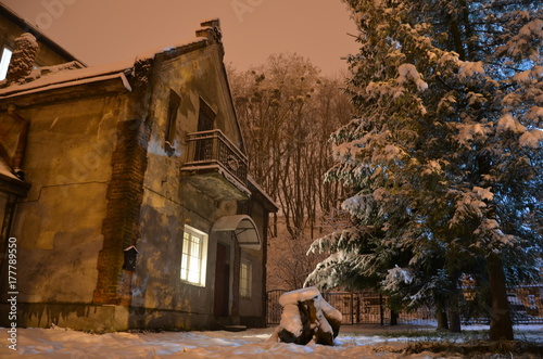 old house with lights from the window at night during the snowy winter next to big christmas tree