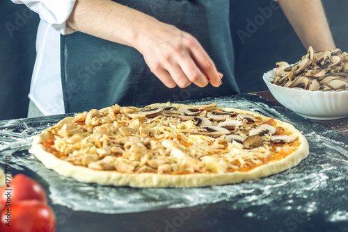 The chef in black apron makes pizza with his hands putting the ingredients for the pizza on the table.