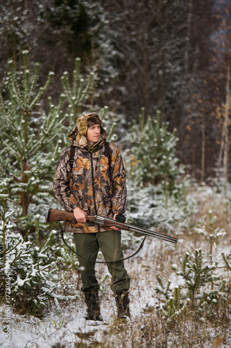 Male hunter with backpack, armed with a rifle, standing in a snowy winter forest.