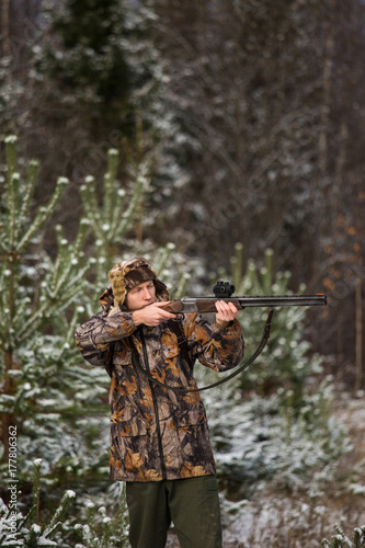 Male hunter in camouflage and with backpack, armed with a rifle, standing in a snowy winter forest.