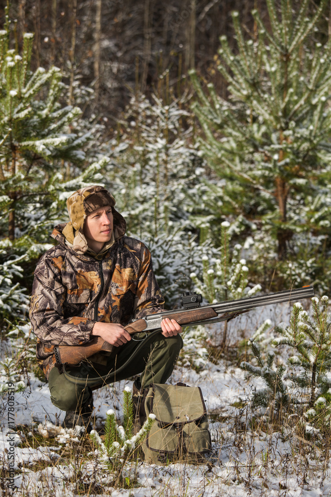 Male hunter in camouflage, armed with a rifle, sitting in a snowy winter forest.  Man is charging a hunting rifle. Winter snow-covered forest.
