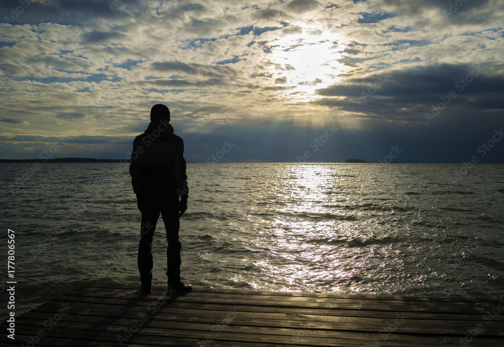 Silhouette of lonely man in jacket and cap who standing on wooden dock and looking on sunset over ocean. Cloudy weather.