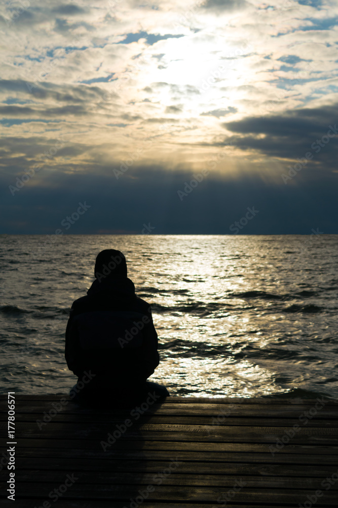 Silhouette of lonely man in jacket and cap who seating on wooden pier and looking on sunset over sea. Cold autumn. Vertical.