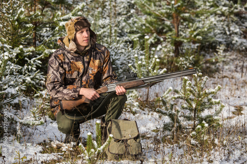 Male hunter in camouflage, armed with a rifle, sitting in a snowy winter forest. Man is charging a hunting rifle. Winter snow-covered forest.