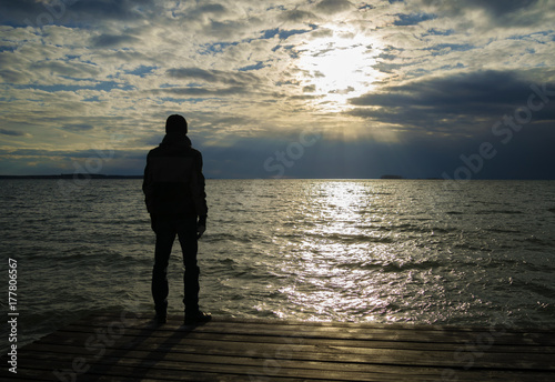 Silhouette of lonely man in jacket and cap who standing on wooden dock and looking on sunset over ocean. Cloudy weather.