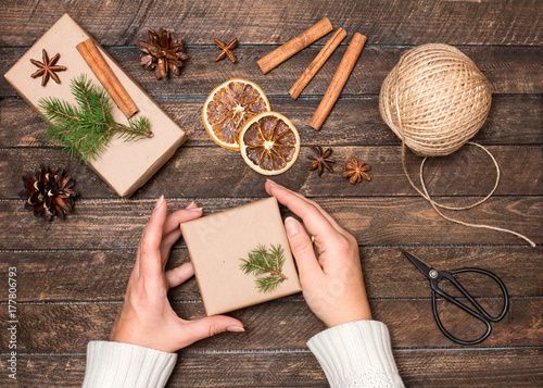 Woman decorating Christmas gifts. Presents wrapping inspirations. Hands, gift boxes, ball of jute, cinnamon sticks, anise, orange slices, fer tree branches, retro scissors on rustic background.