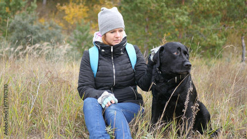 A girl sits on a yellow grass and holds her dog by the collar. Photo
