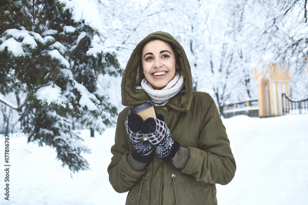 Obraz premium young beautiful brunette woman in a warm jacket with hoodie and warm mittens is walking outdoors in winter. a woman holding a paper glass with hot tea or coffee. Enjoys a walk on a frosty day