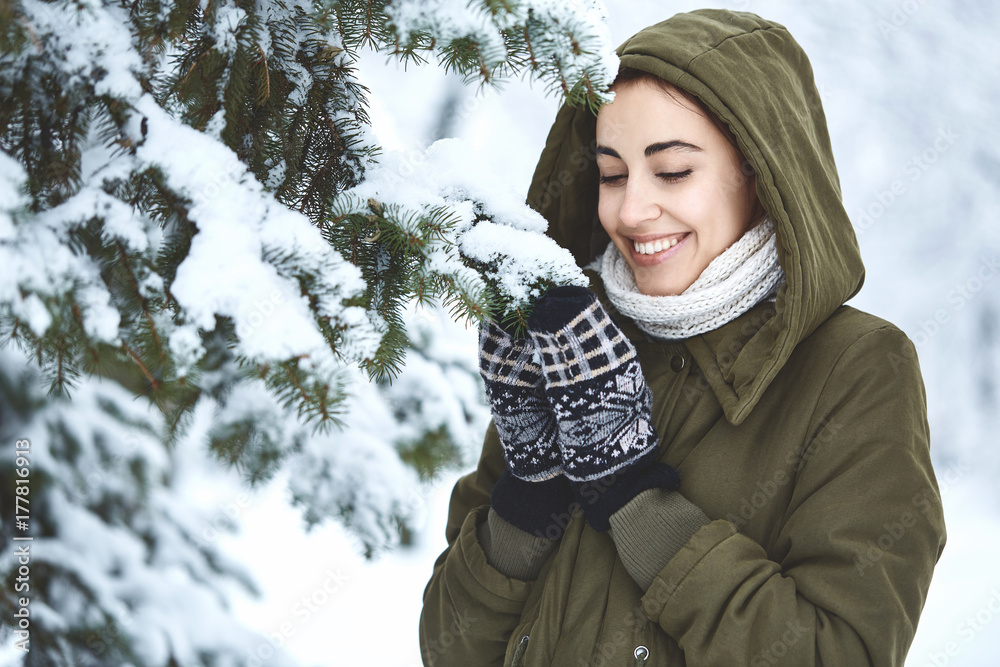 young beautiful brunette woman in a warm jacket with hoodie and warm mittens is walking outdoors in winter. a woman stands under the fir branches with snow and play with them young beautiful brunette woman in a warm jacket with hoodie and warm mittens is walking outdoors in winter. a woman stands under the fir branches with snow and play with them