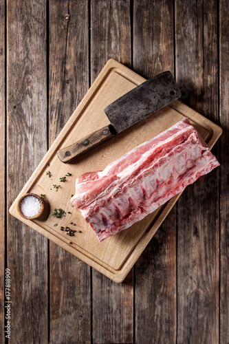 Fresh raw uncooked whole rack of pork loin with ribs on wooden cutting board with salt, thyme and butcher clever over old wood plank table. Top view, space.
