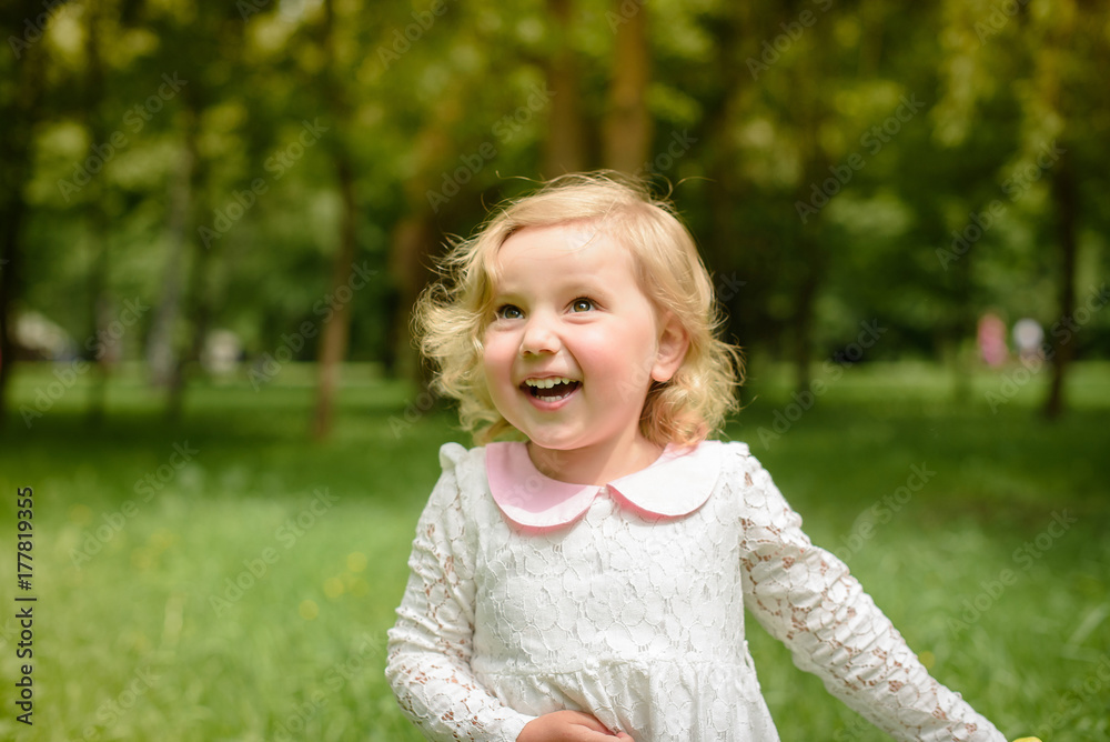 Beautiful little blonde girl, has happy fun cheerful smiling face, white dress. Portrait nature.