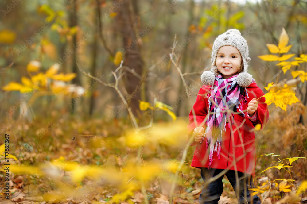 Cute little girl having fun on beautiful autumn day. Happy child playing in autumn park. Kid gathering yellow fall foliage.