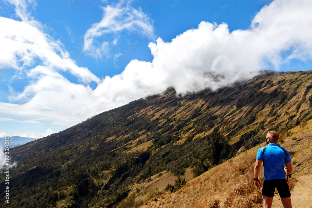 Sporty guy traveler in a blue t-shirt on a background of a beautiful sky with clouds in the mountains. Trekking to Rinjani Volcano. Indonesia. Lombok Island.