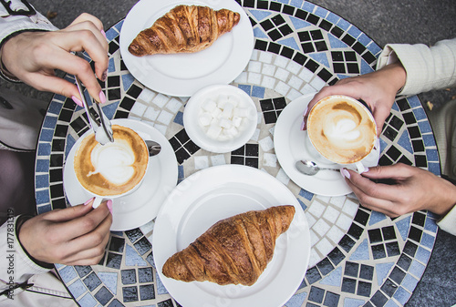 Top view of two women having coffee brake in French cafe. Women friends drinking coffee in French styled restaurant. Croissants with coffee on vintage table. Girls enjoying coffee outdoors.