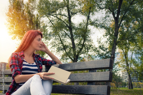 Thoughtful redhead woman relaxing on park bench with a book.