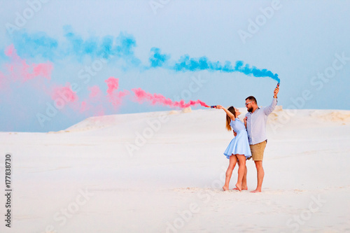 young couple  standing on a sand and holding smoke bomb and looking at each other, romantic couple with blue color and red color smoke bomb on beach