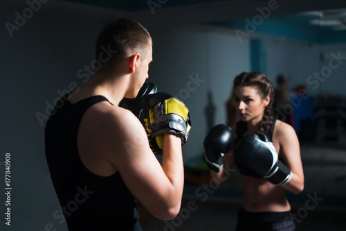Young fighter boxer fit girl wearing boxing gloves in training with personal trainer in gym. Low key image. Moment before punch