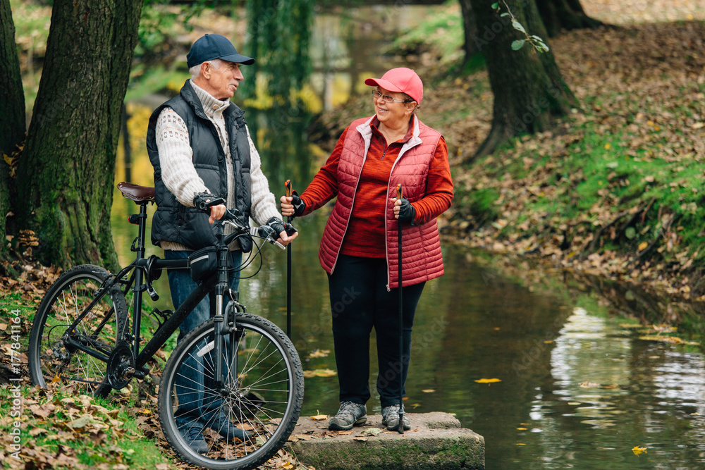Pretty senior sport couple standing in colorful autumn park near river. Mature woman with nordic walking poles and old man with bicycle resting outdoors.