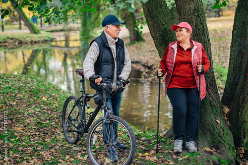 Pretty senior sport couple standing in colorful autumn park near river. Mature woman with nordic walking poles and old man with bicycle resting outdoors.