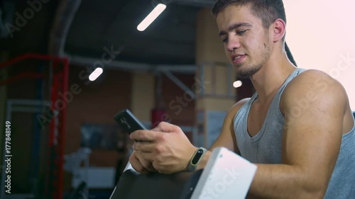 Athletic man sitting in fitness gym after workout, chatting online smart phone