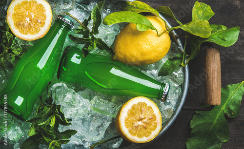 Bottles of green lemonade on chipped ice in metal tray with fresh lemons and mint over dark wooden background, top view